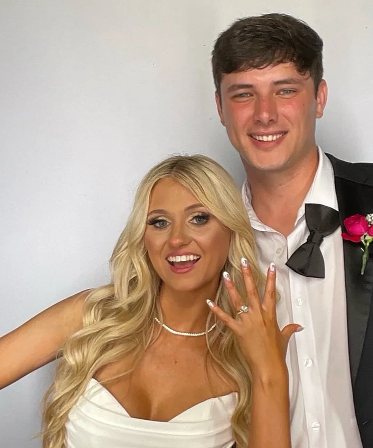 Woman in a white strapless dress shows her ring beside a man in a bow tie and boutonniere