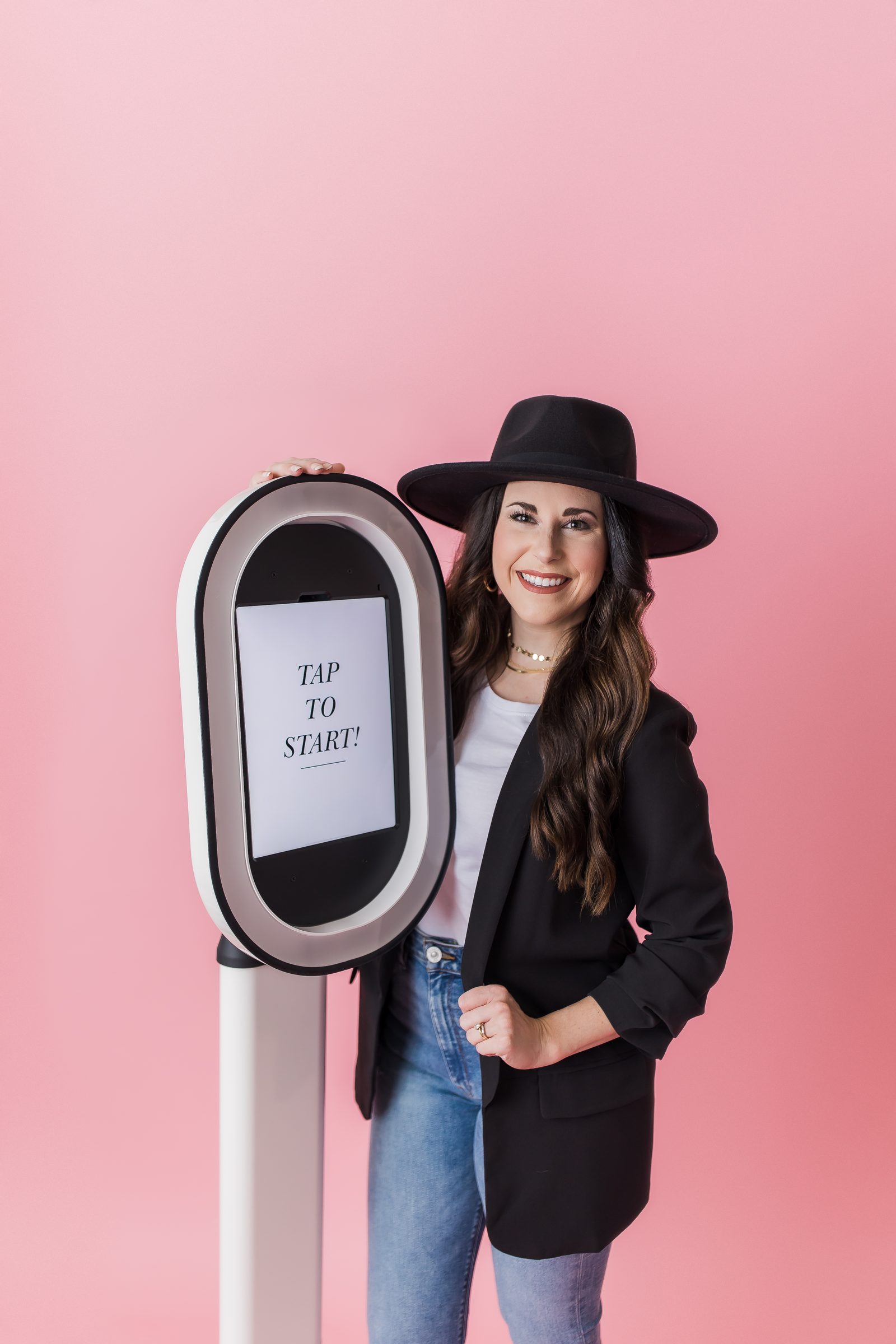 Woman in a black hat and blazer beside a white photo booth reading Tap to start, pink backdrop