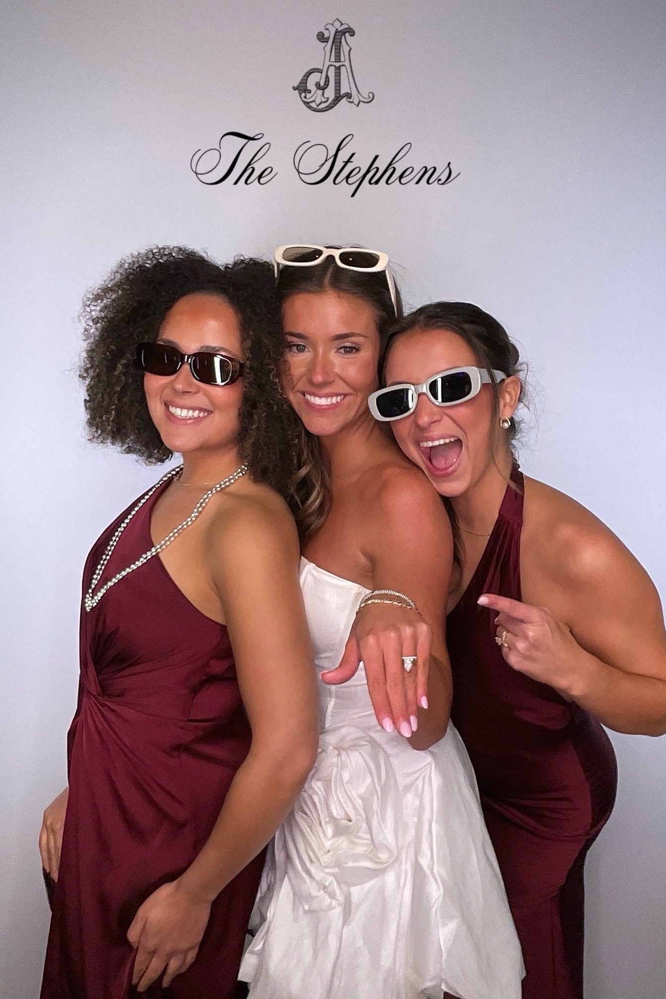 Woman in a white dress shows her ring between two women in burgundy dresses and sunglasses